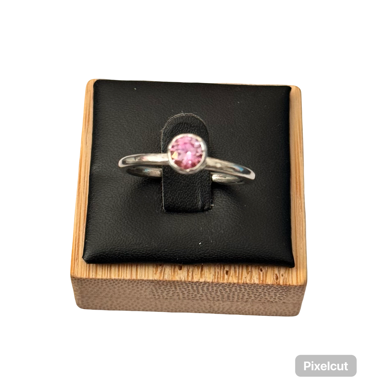 A silver ring with a round pink gemstone displayed on a black velvet stand inside a light wood box, set against a plain white background. The stone is a certified moissanite and metal is sterling silver