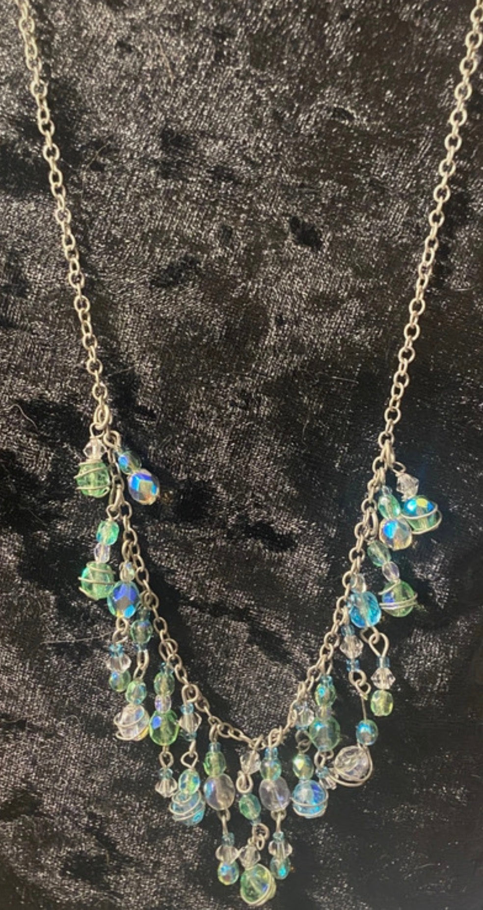 A silver-toned beaded necklace with iridescent blue, green, and clear faceted beads, displayed against a dark textured fabric background.