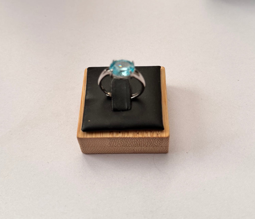 A sterling silver hallmarked ring with a round-cut light blue gemstone displayed on a black cushioned wooden jewellery box against a plain light grey background.