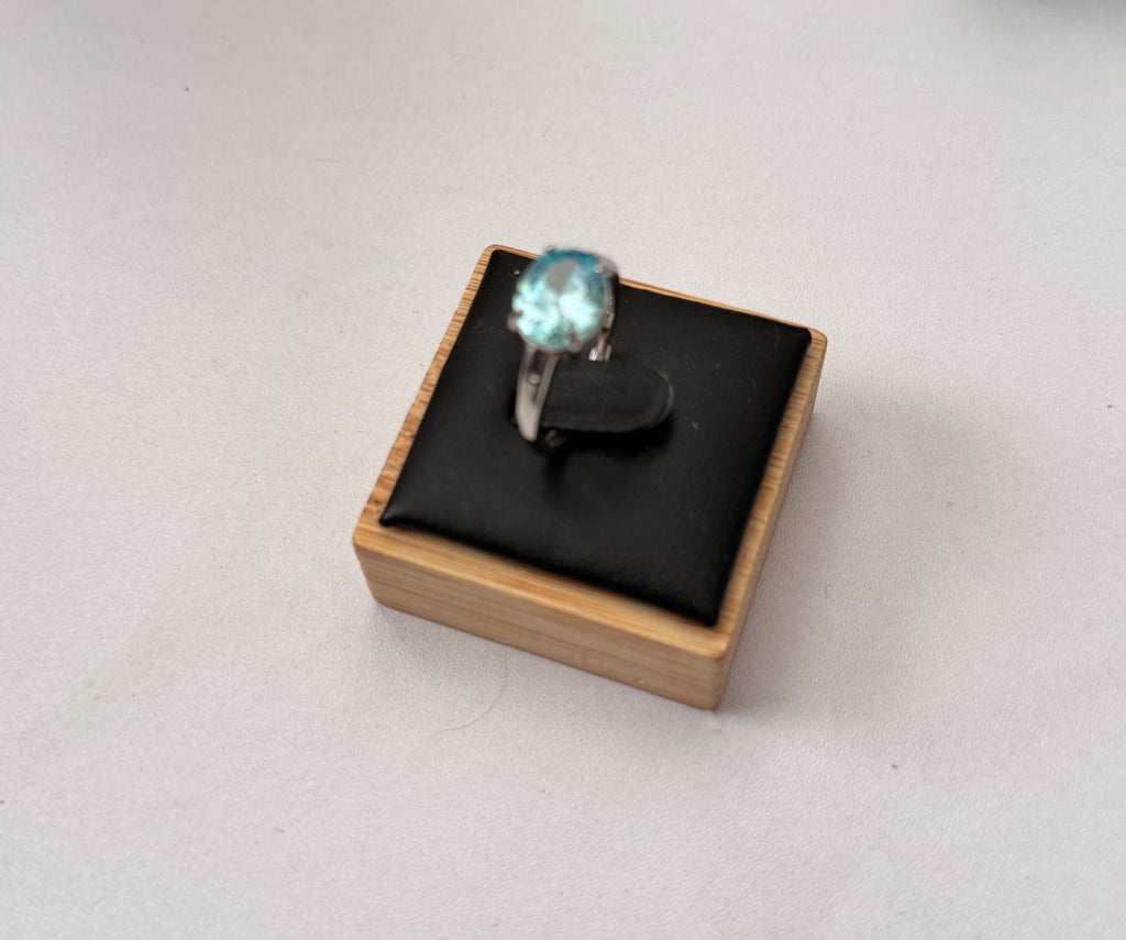 A silver ring with a round light blue gemstone displayed on a small wooden jewelry box with a black cushion, set against a plain light gray background.
