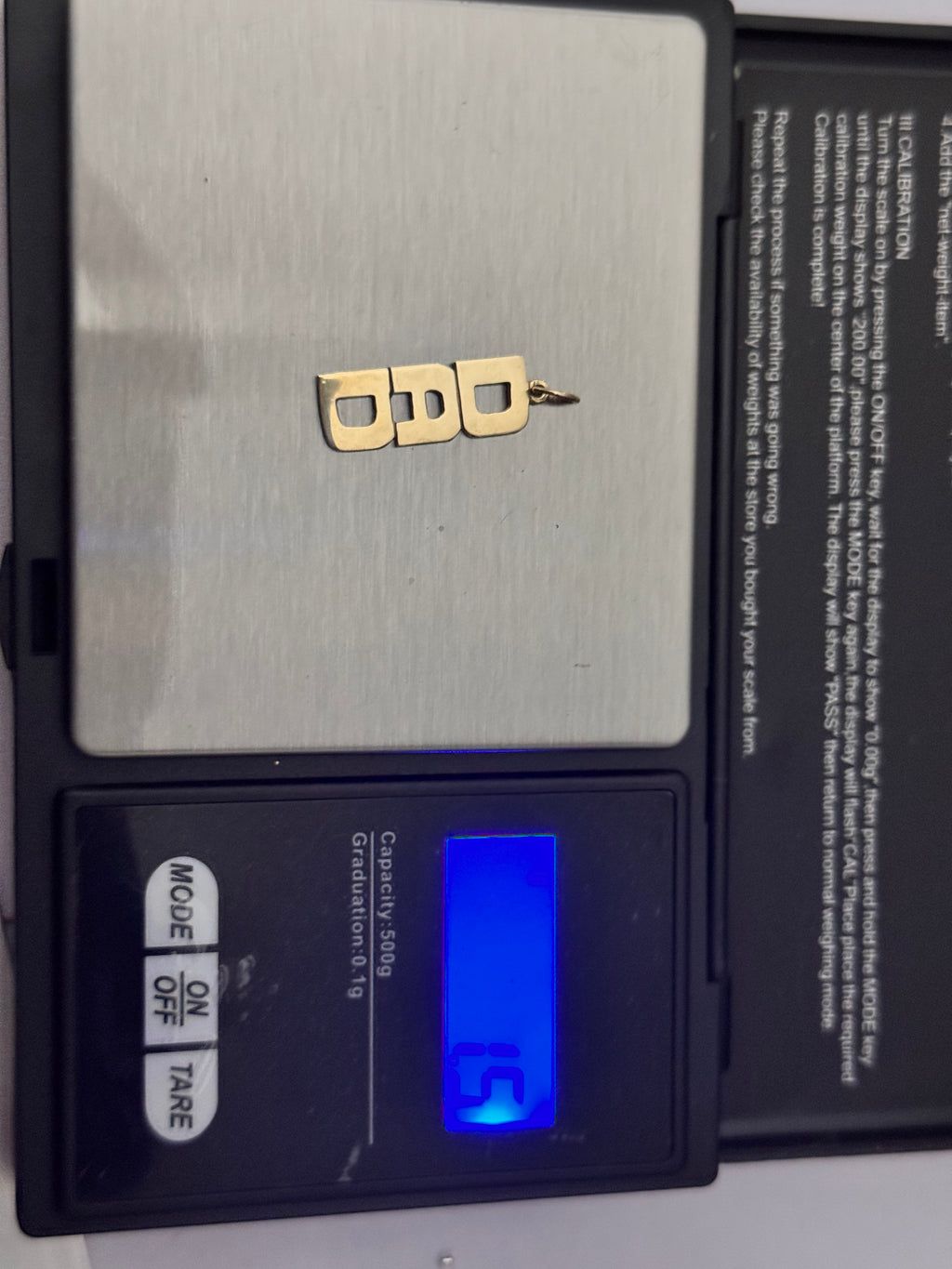 A small 9 ct gold pendant spelling 'DAD' in block letters is shown on a digital pocket scale with a silver weighing platform and a black display screen showing 1.5 grams.