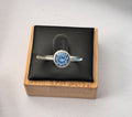 A sterling silver ring with an uncertificated lab sapphire a round gemstone, displayed on a black cushion inside a wooden box.