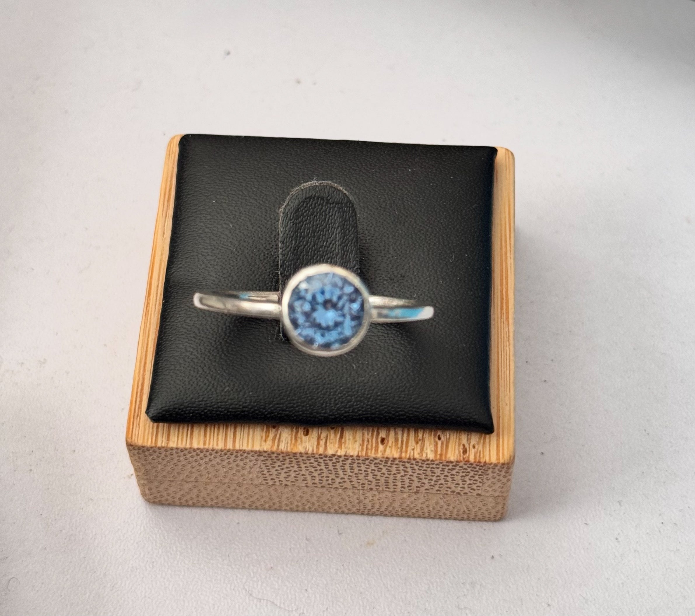 A sterling silver ring with an uncertificated lab sapphire a round gemstone, displayed on a black cushion inside a wooden box.