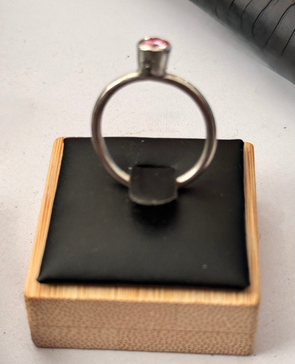 A Aterling silver ring with a 5mm round pink moissanite gemstone, displayed on a black cushion atop a light wood box.
The Moissanite is certified