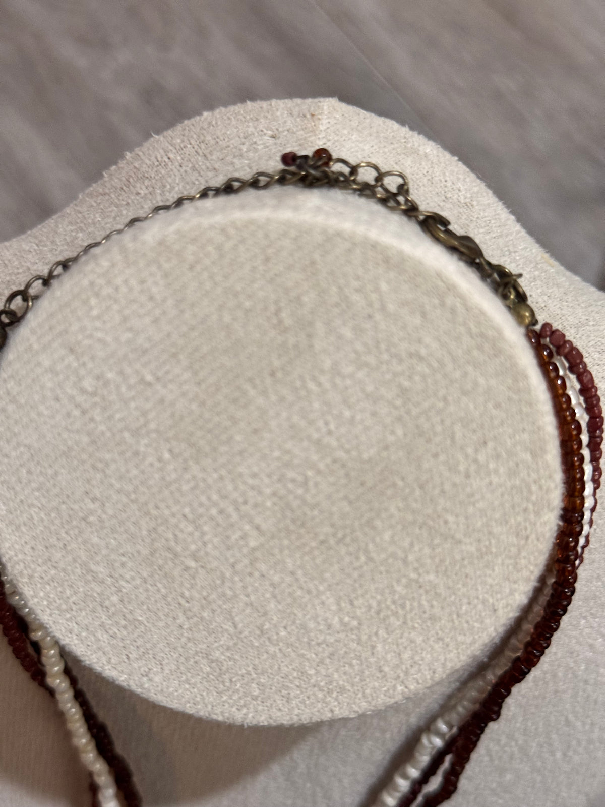 A close-up of a multi-strand necklace displayed on a round cream-colored jewelry bust. The necklace features a dark bronze chain, strands of dark brown translucent beads, and strands of light beige/cream beads.