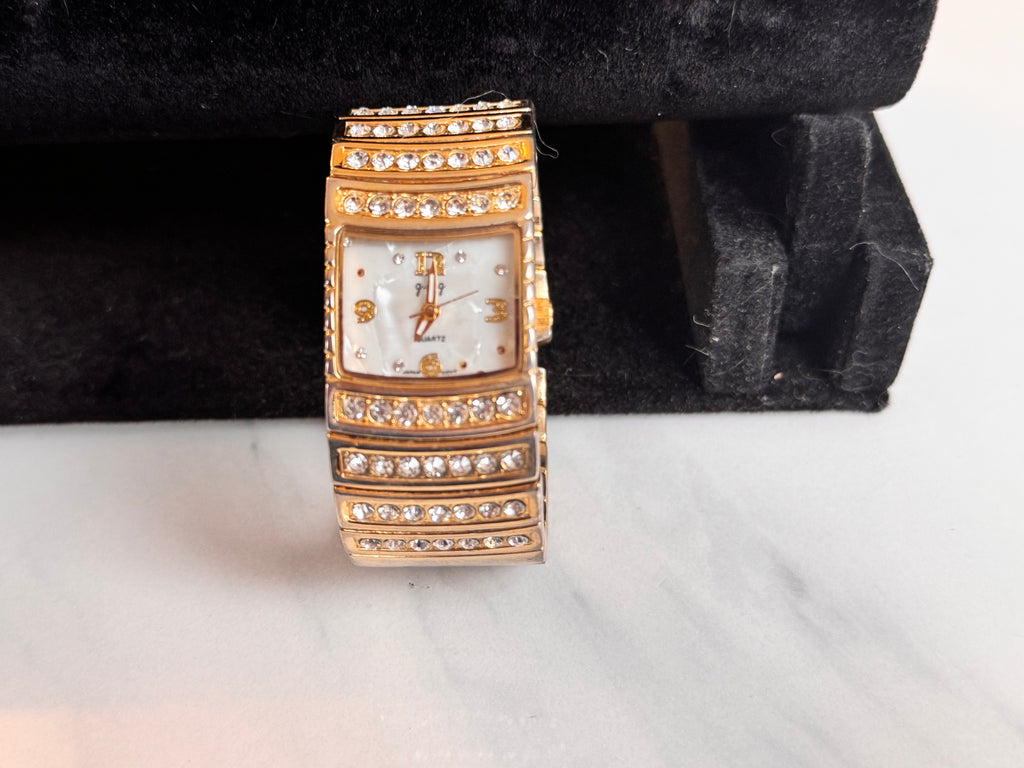 A gold-tone rectangular watch with a mother of pearl dial and multiple rows of clear rhinestones on the case and band, displayed upright on a black velvet jewelry stand over a light marble surface. Full working order