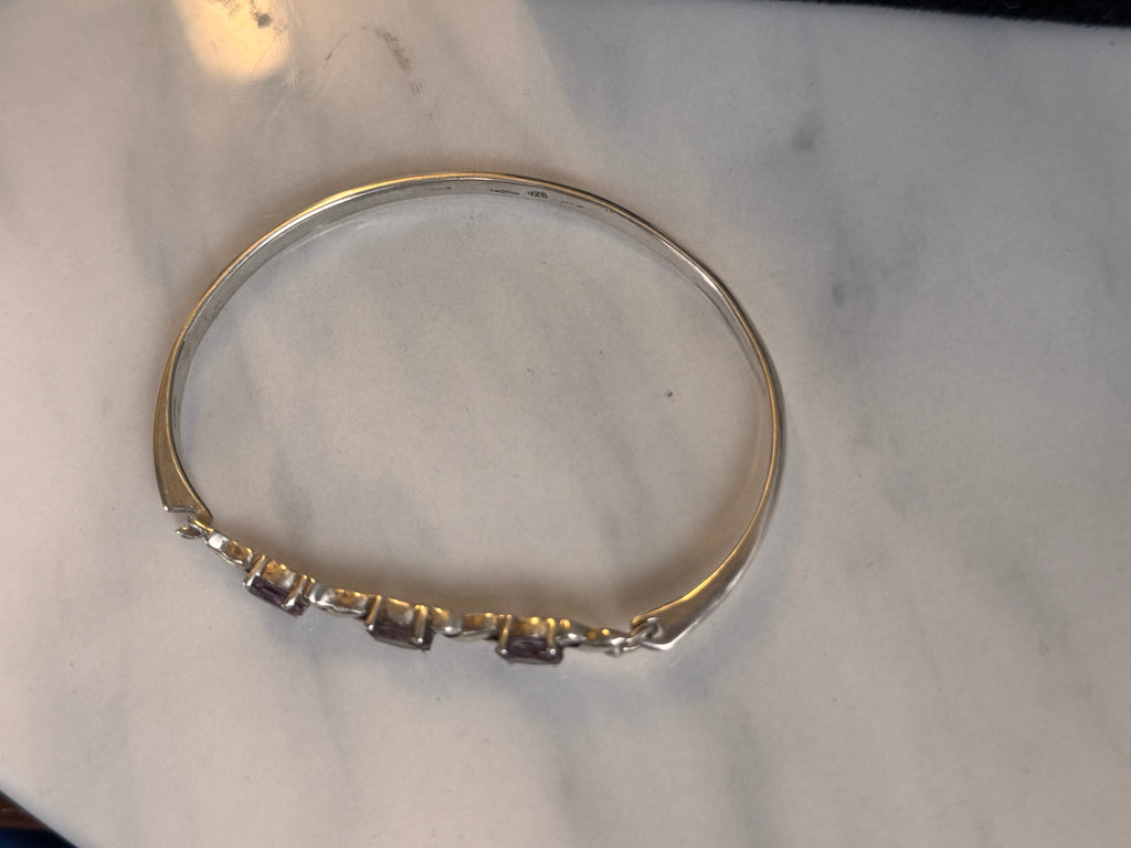 A silver hallmarked bangle bracelet with three small oval-cut purple gemstones set along the front band, displayed on a light marble surface.