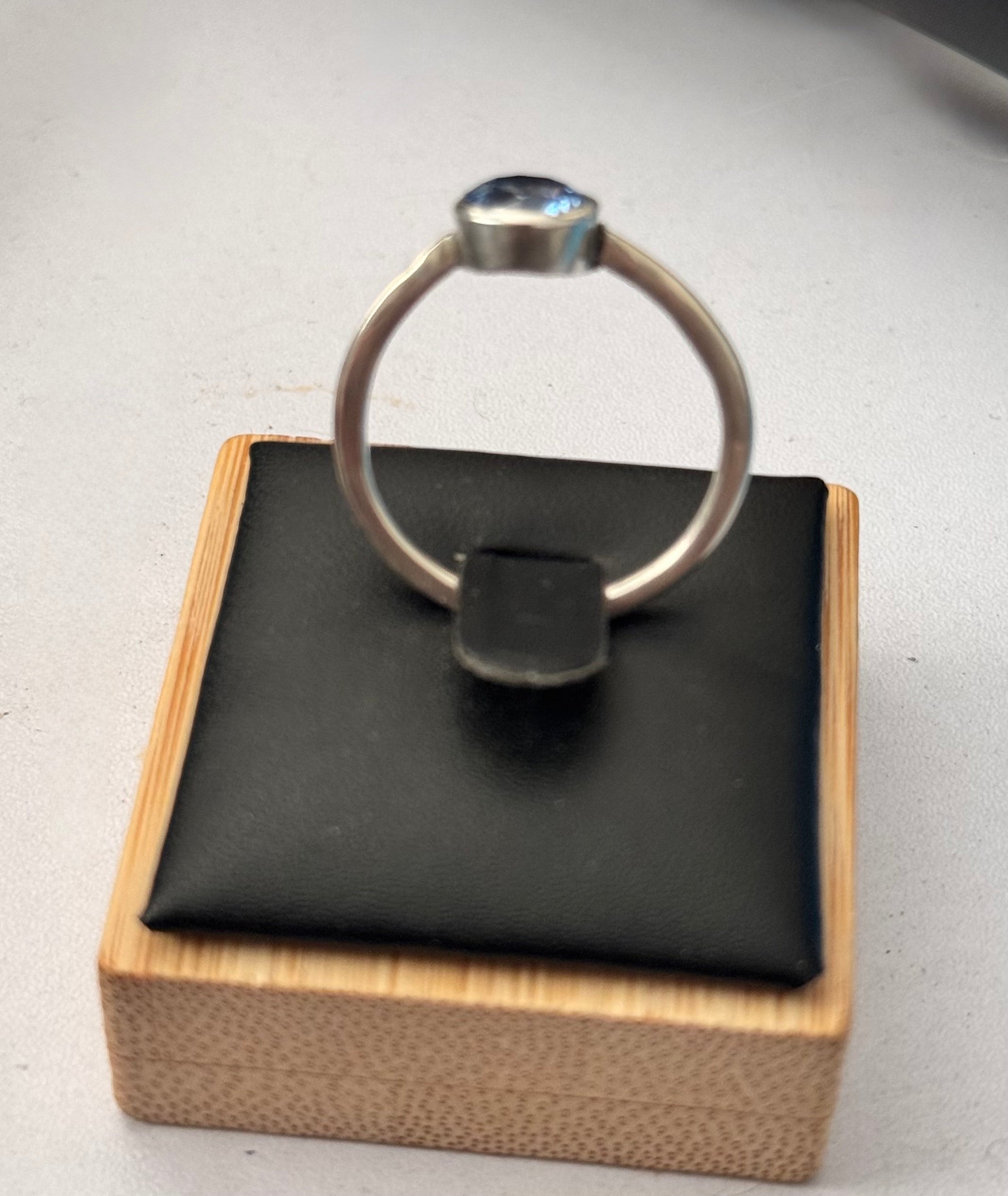 A silver ring with a round bezel-set Mid blue lab sapphire gemstone, displayed on a black cushion inside a light wood box.