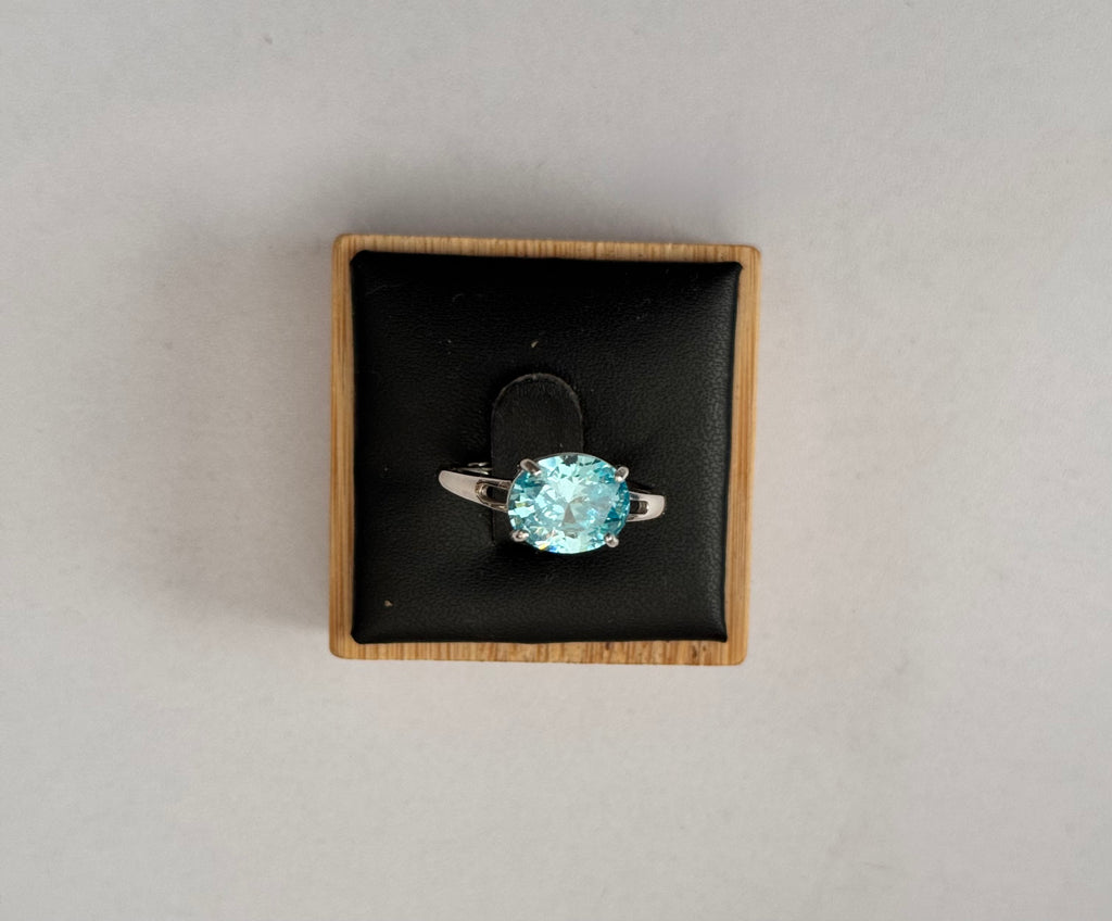 An oval-cut light blue gemstone ring set in a silver-colored band, presented in a square black velvet jewelry box with a light wood border, shot from a top-down view on a neutral light gray background.