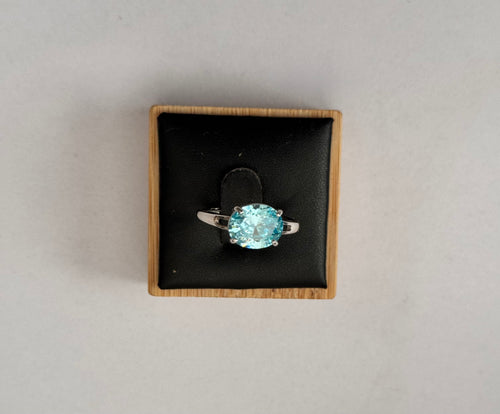 An oval-cut light blue gemstone ring set in a silver-colored band, presented in a square black velvet jewelry box with a light wood border, shot from a top-down view on a neutral light gray background.