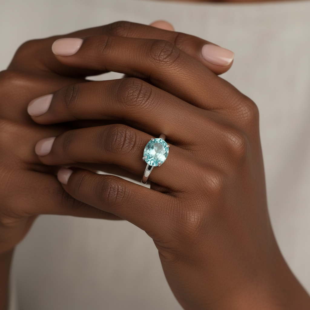 A close-up of a person's hands wearing a silver ring with a large oval light blue gemstone, set against a soft beige background. This is an Ai Projection of the ring on a person so give an idea of what it will look like on the ring finger