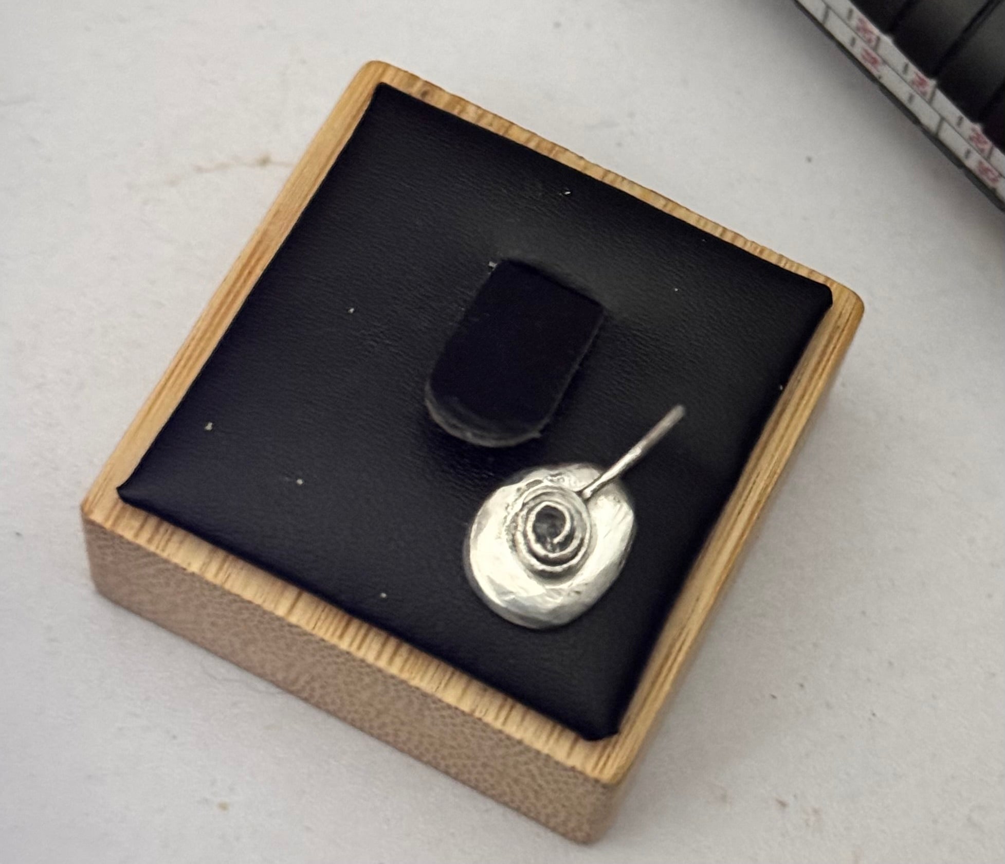 A sterling silver oval piece with a textured hammered finish and a simple rose spiral in the centre of which ashes can be added and sealed with resin, displayed on a black wooden jewelry display on a light gray surface.