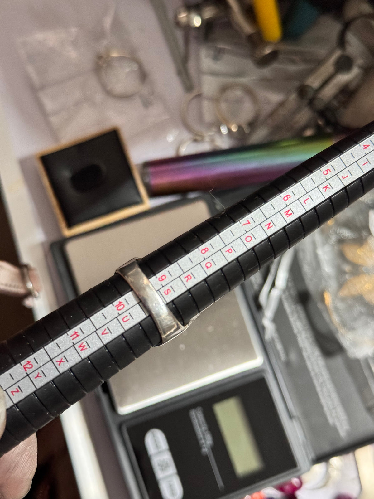 A black ring madrel showing the thick  band on a ring mandrel showing it is size T held in a hand over a scale and jewelry accessories on a workshop table.