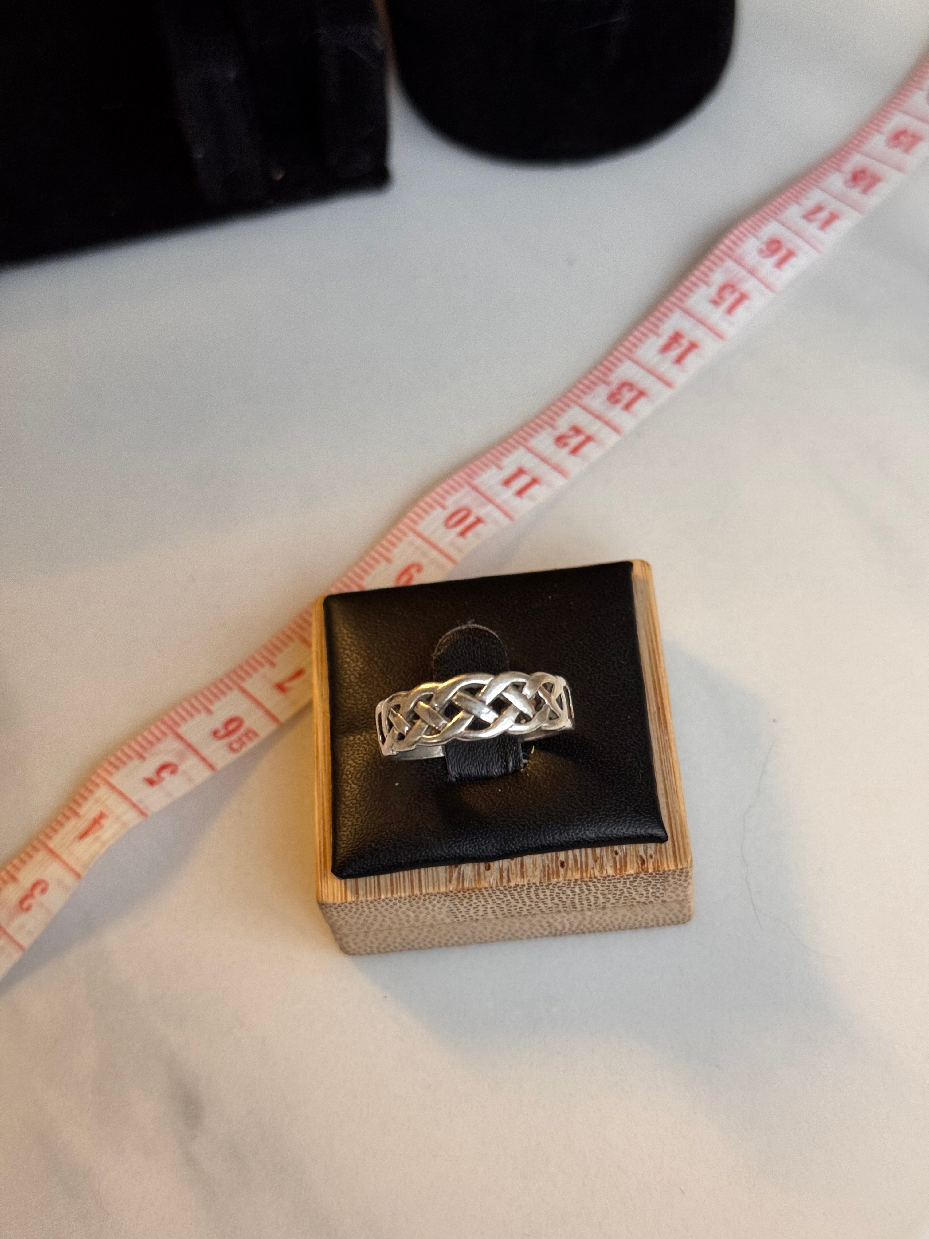 A sterling silver hallmarked ring with a Celtic knot design displayed on a black ring box set in a wooden base, placed on a white marble surface next to a measuring tape.