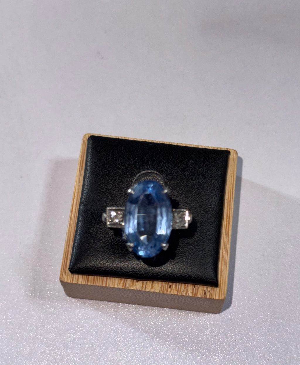 An oval-cut blue gemstone ring, possibly paste, with silver colour, 950 Palladium. 2 side stones, diamonds, displayed on a black cushioned wooden jewelry ring display against a light gray background.