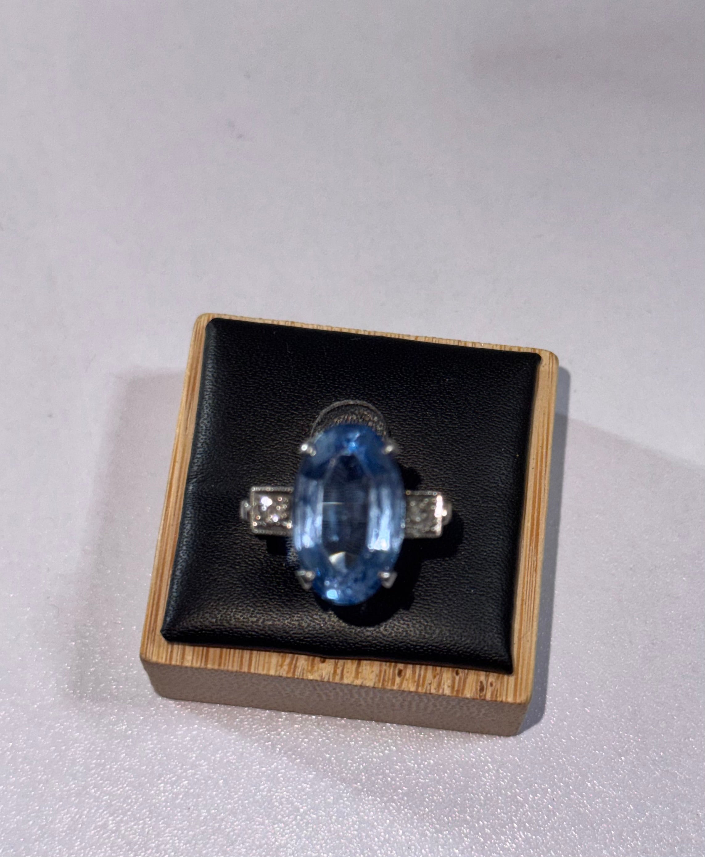 An oval-cut blue gemstone ring, possibly paste, with silver colour, 950 Palladium. 2 side stones, diamonds, displayed on a black cushioned wooden jewelry ring display against a light gray background.
