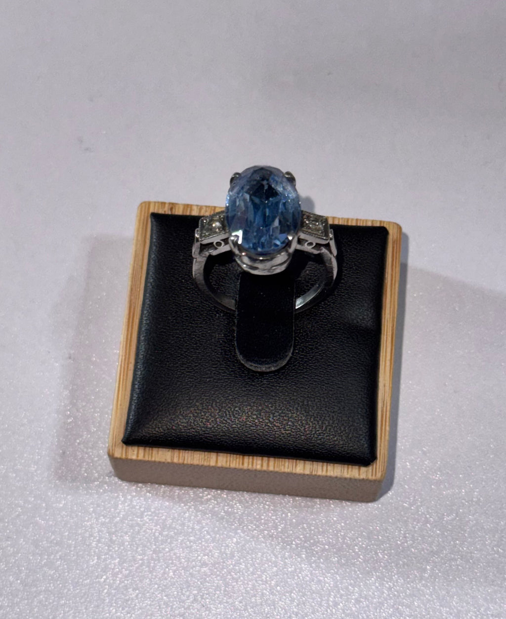 A Palladium 950 ring with a large oval blue gemstone & 2 diamonds, displayed on a black cushion inside a light wood box against a light gray background.
