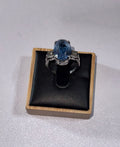 A Palladium 950 ring with a large oval blue gemstone & 2 diamonds, displayed on a black cushion inside a light wood box against a light gray background.