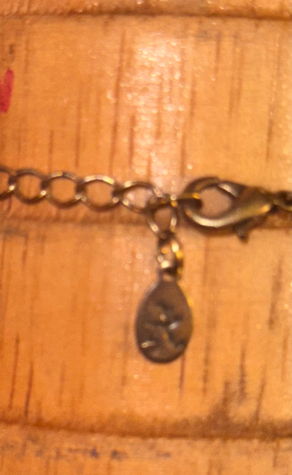 Close-up of a gold-toned jewellery tag showing the Accessorize symbol on a small teardrop charm, lobster clasp and  resting on a warm wood surface.