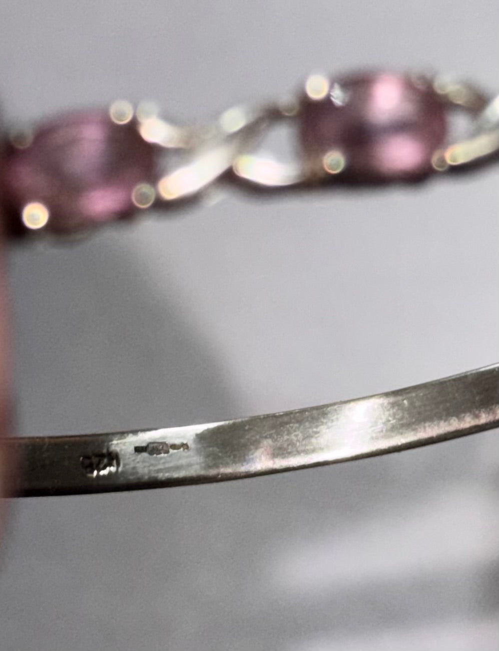 Close-up of a silver-toned metal bracelet showing a polished band with a stamped hallmark and part of The decorative front with oval pinkish-purple amethyst stones in the background.