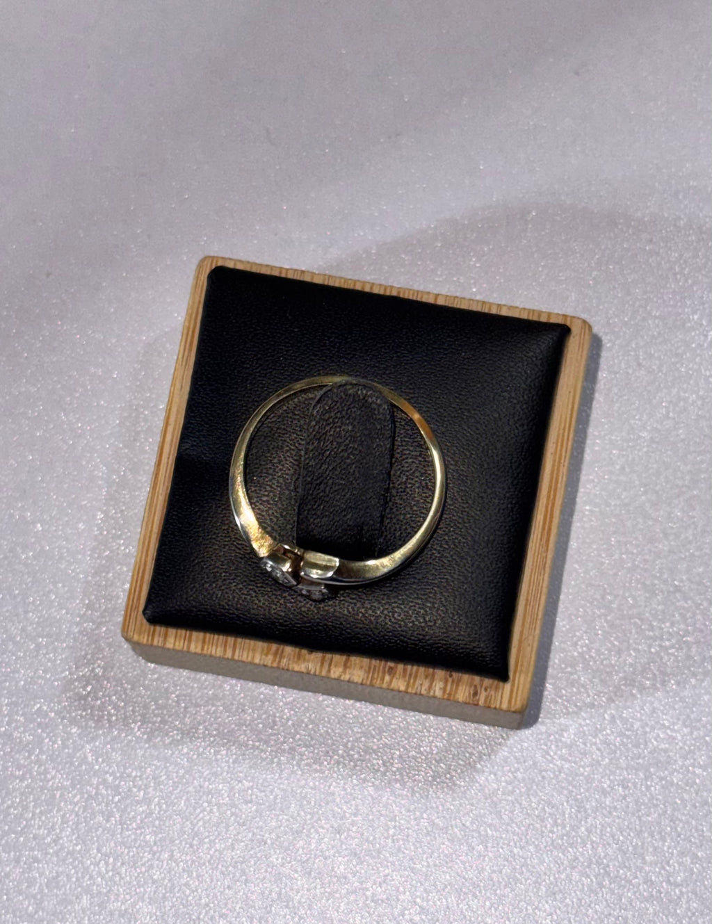 A gold 18ct ring displayed on a black cushion inside a square wooden presentation box, set against a light textured background.