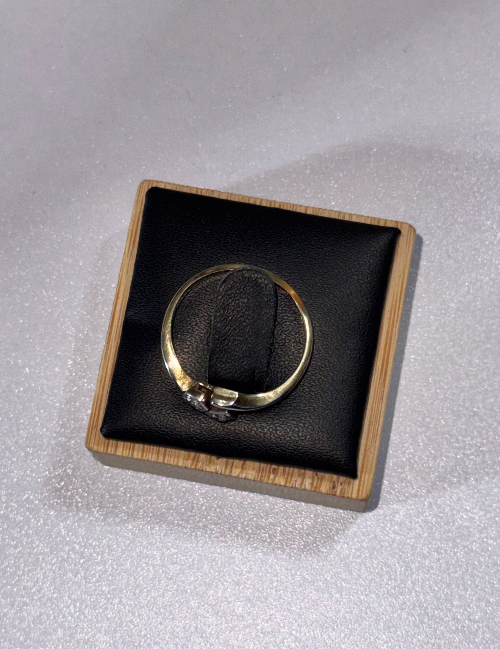 A gold 18ct ring displayed on a black cushion inside a square wooden presentation box, set against a light textured background.
