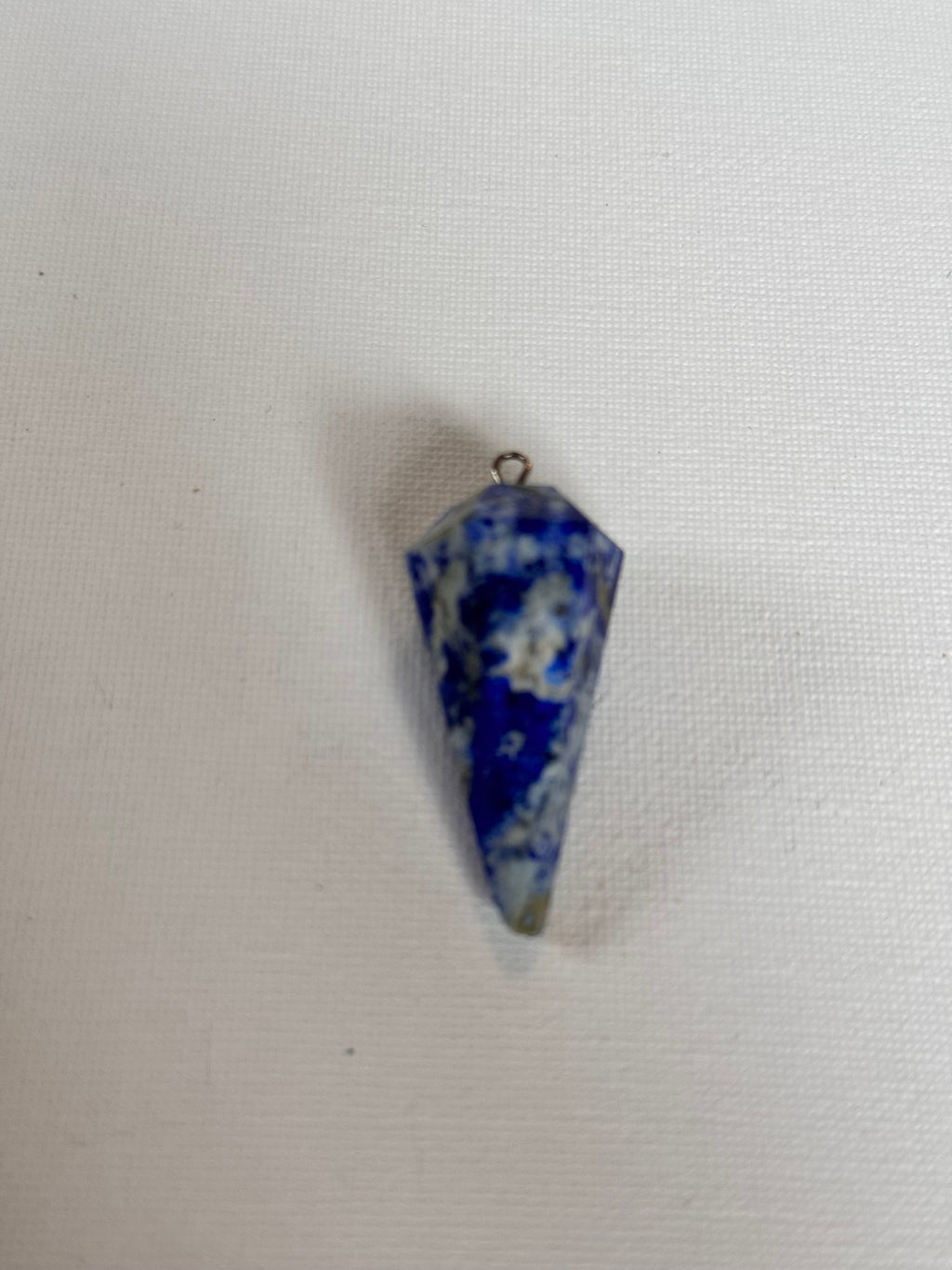 A small pointed blue and white marbled stone pendant with a tiny metal loop with a flower at the top, placed on a white textured fabric background.