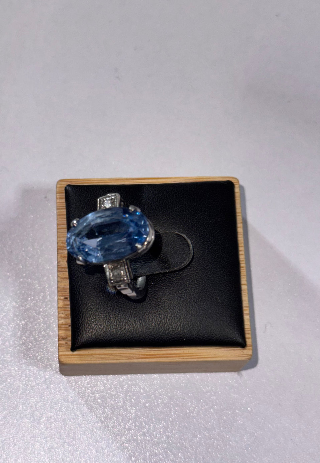 A silver ring (950 Palladium) featuring a large oval-cut light blue gemstone ( possibly paste ) and small clear accent stones, (diamonds) displayed on a black cushioned square  ring display on a light wood border.