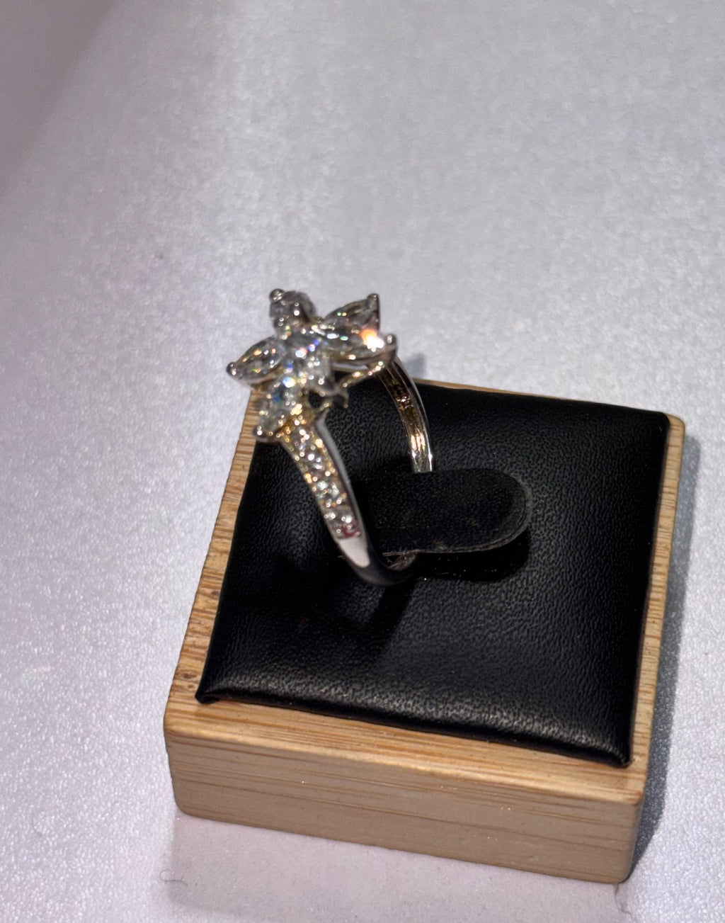 A side view of the sterling Silver Starburst Ring with a star-shaped cluster of clear stones, displayed on a black cushion inside a light wood box against a light gray background.