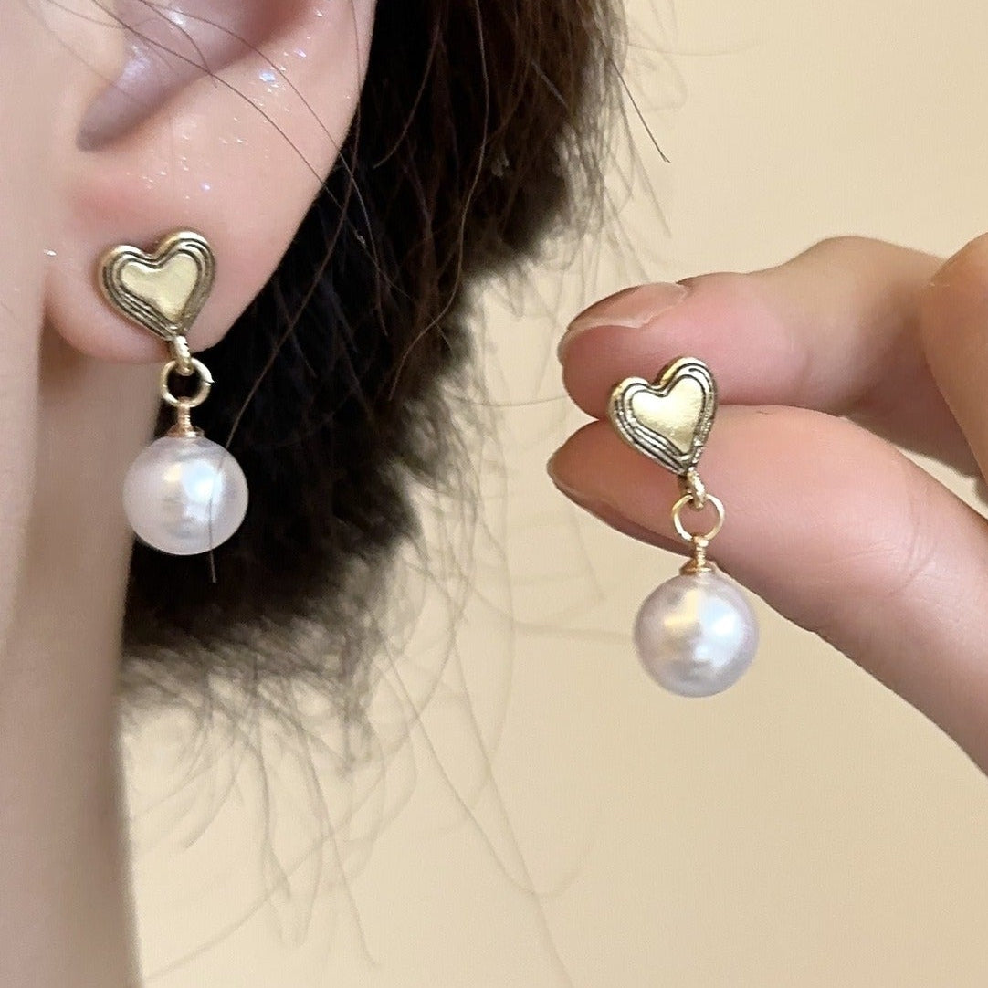 A close up of the earrings one in an ear and the other in her fingers. very pretty faux pearl and heart stud earrings