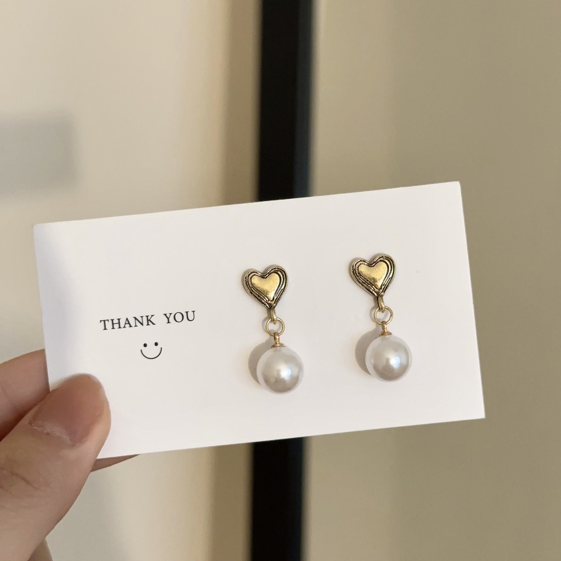 Another image angle of the Heart and faux peal earrings on a white Card Background with a smiley face and a thank you, being held in finger and thumb