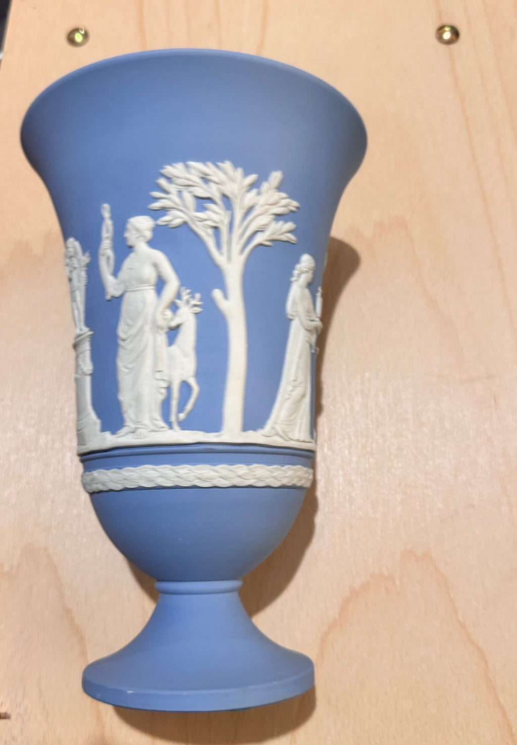 A blue and white Wedgwood pedestal vase with classical figures and a tree relief, photographed against a light wood background. Vintage Blue Jasperware
