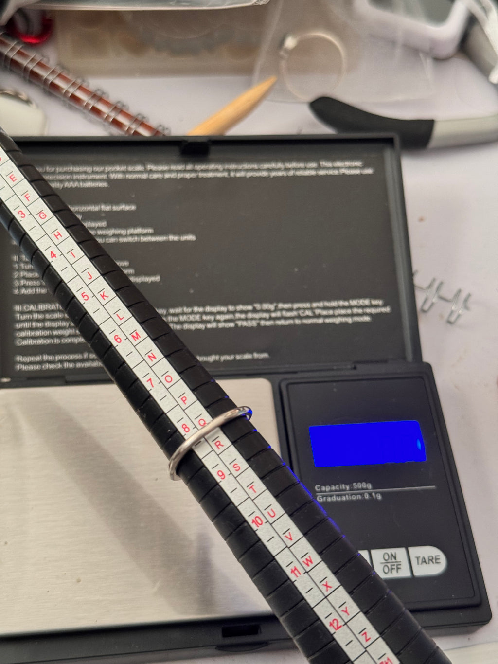 A black plastic ring sizer gauge displaying the ring size its just over a Q. Positioned over a digital scale in a workspace setting.