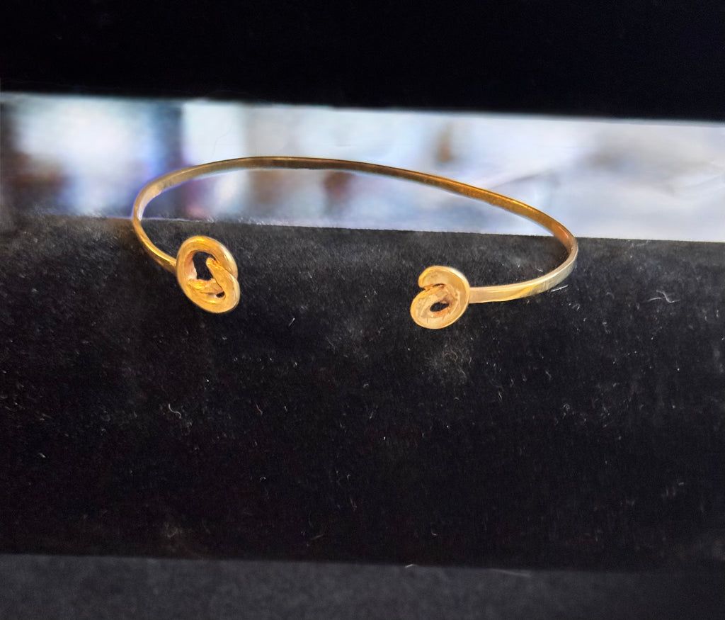 A thin gold cuff bracelet with two open spiral ends, displayed on a black velvet jewelry stand.