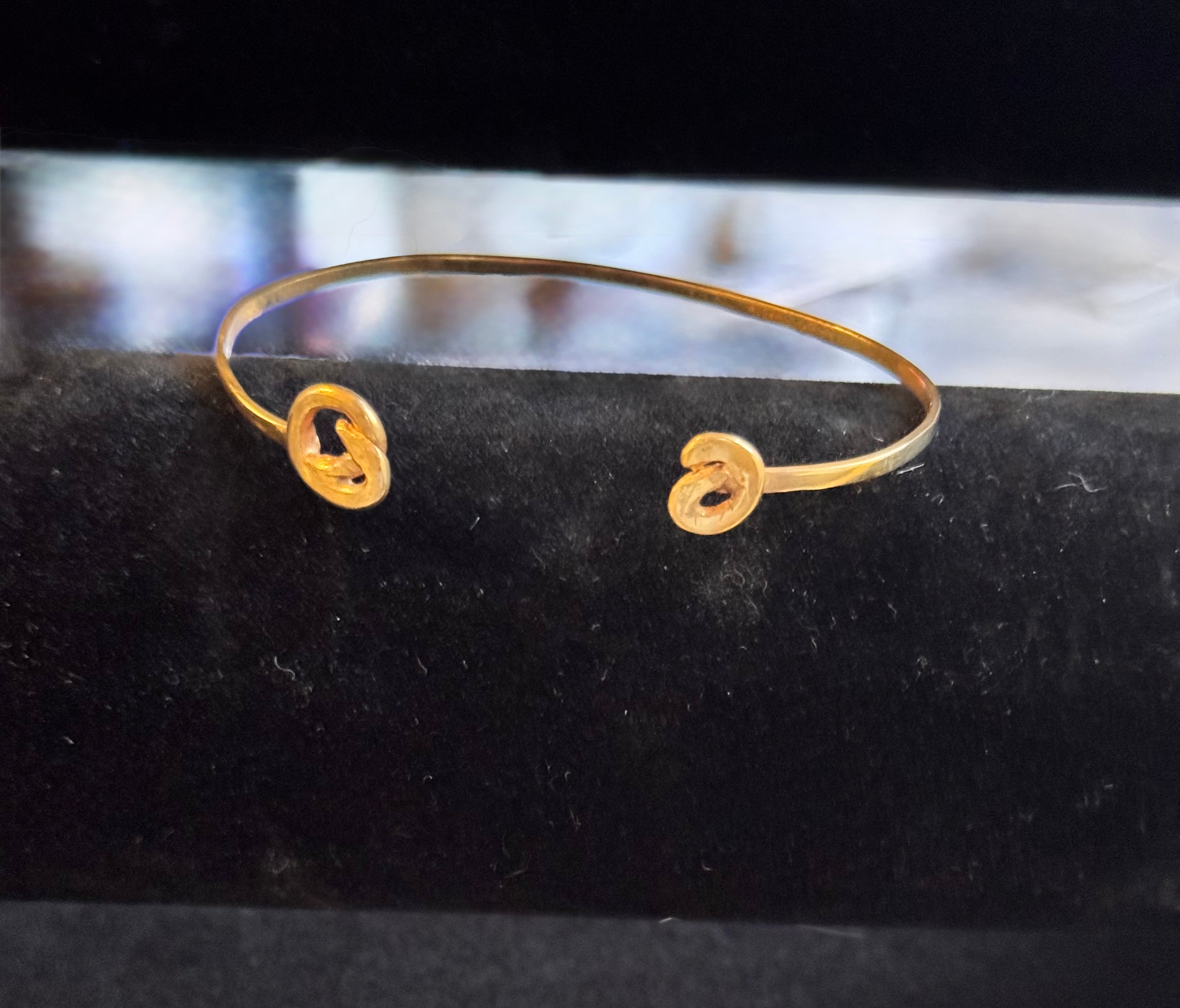 A thin gold cuff bracelet with two open spiral ends, displayed on a black velvet jewelry stand.