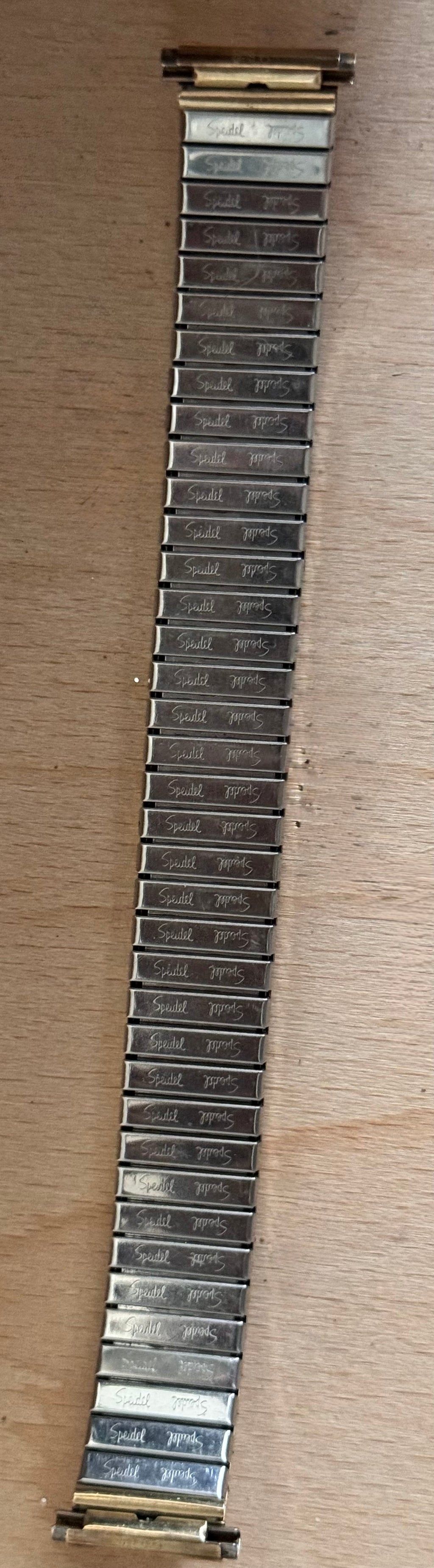 A close-up of a silver-toned metal link watch band with gold-toned end caps, laid flat on a light wood surface.