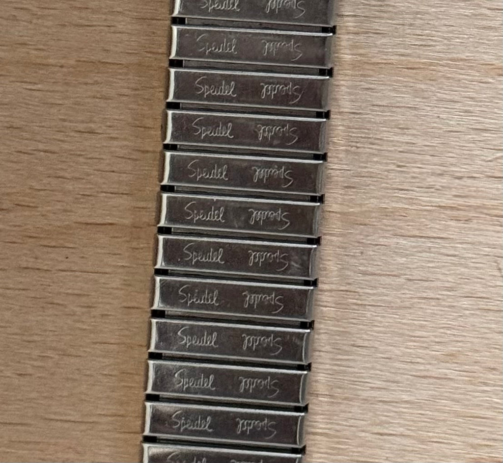 A close-up of a metal bracelet or belt strap laid flat on a light wood surface, with repeated engraved text reading 'Speidel'.