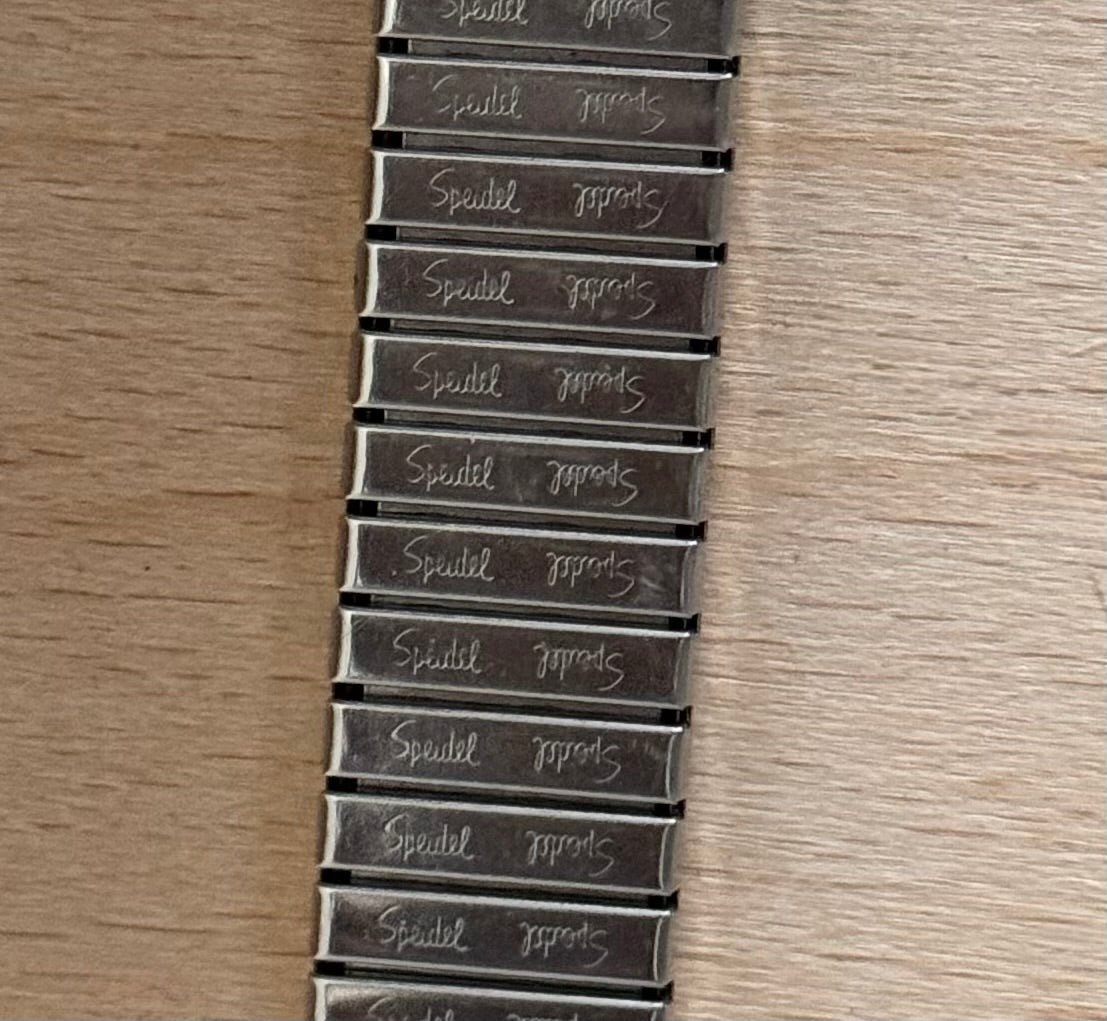 A close-up of a metal bracelet or belt strap laid flat on a light wood surface, with repeated engraved text reading 'Speidel'.