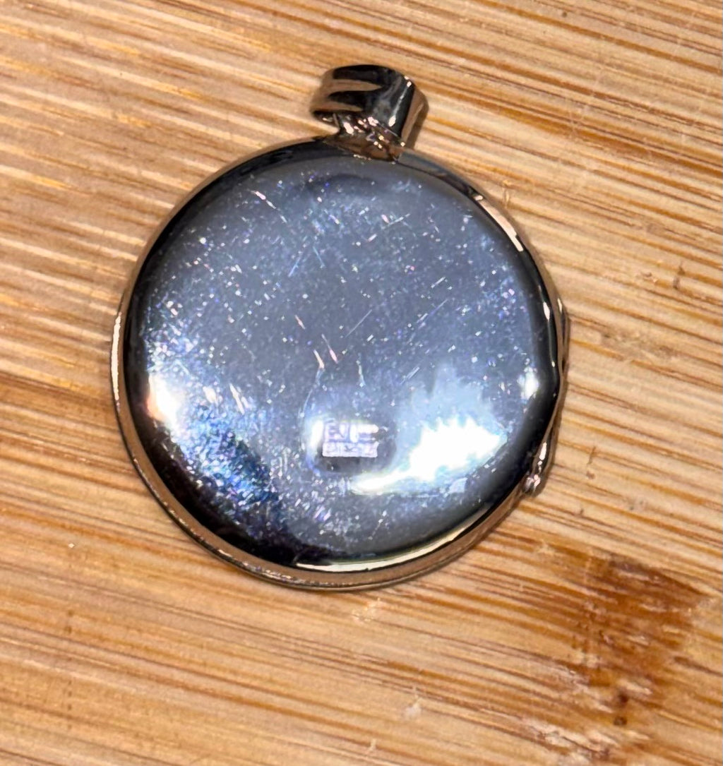 A round silver locket pendant reverse side showing Hallmark, with a polished metallic finish, resting on a light brown wooden surface.