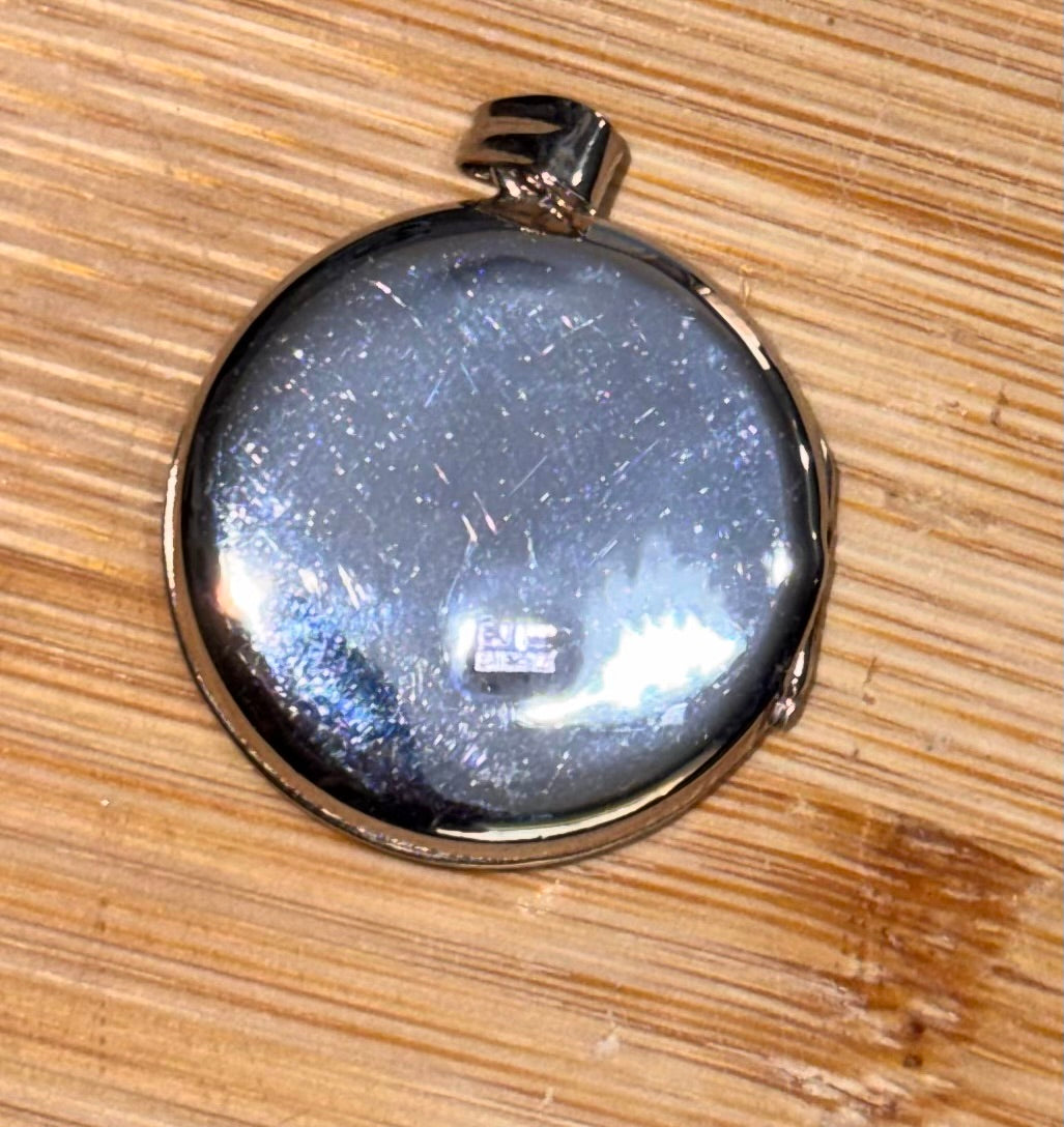 A round silver locket pendant reverse side showing Hallmark, with a polished metallic finish, resting on a light brown wooden surface.