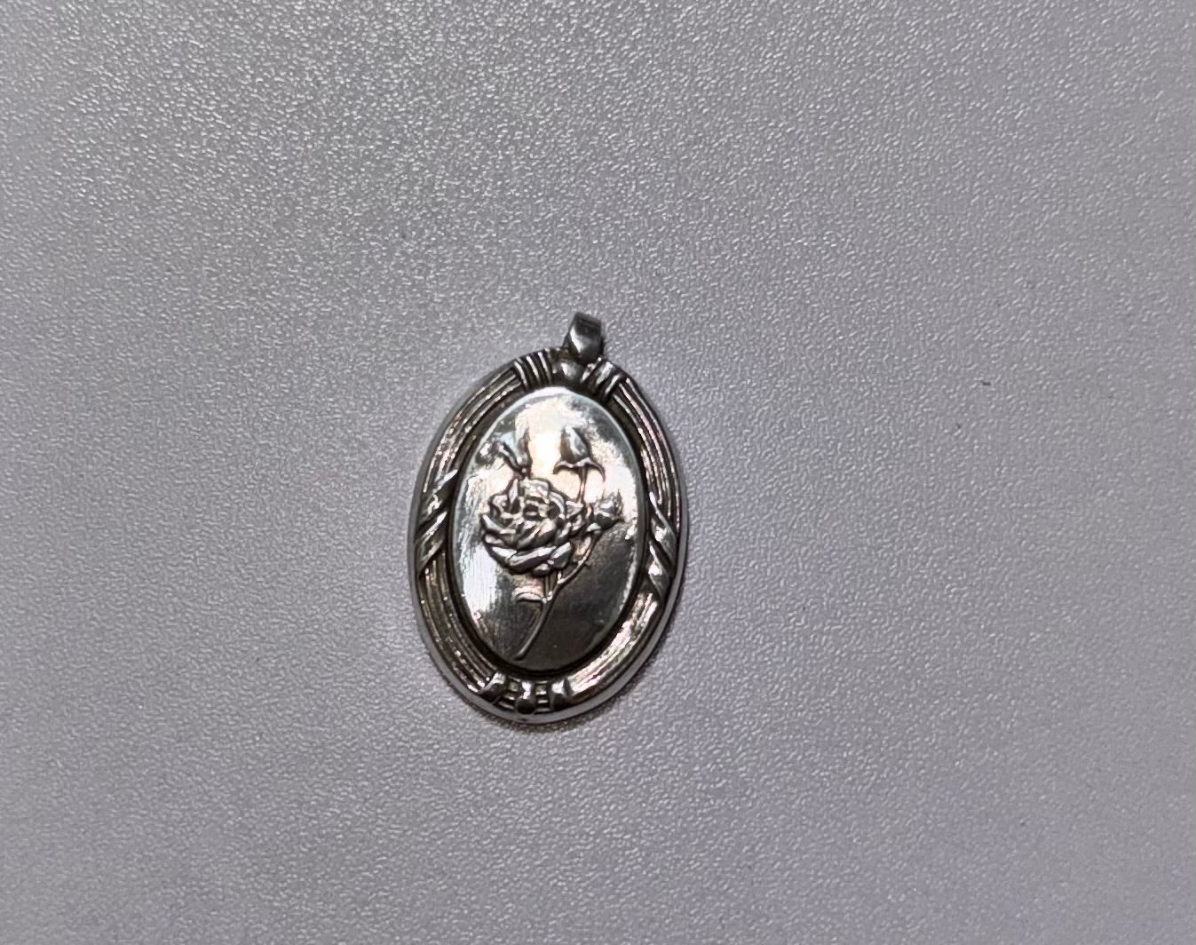 A 2 cm high oval silver pendant featuring an embossed rose design, displayed on a textured gray background.
