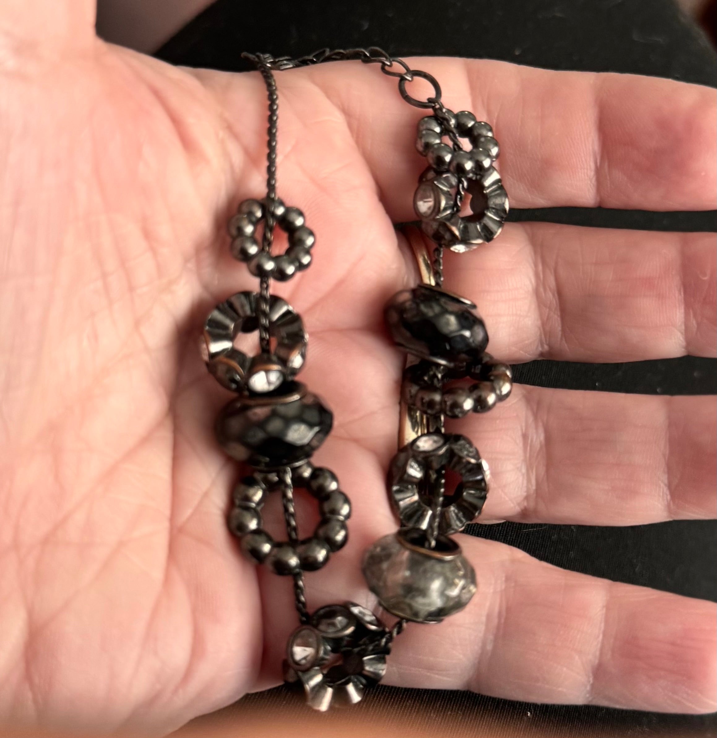 A close-up of a hand holding a dark metal beaded charm bracelet with faceted beads and textured circular charms also in Black, gothic style.