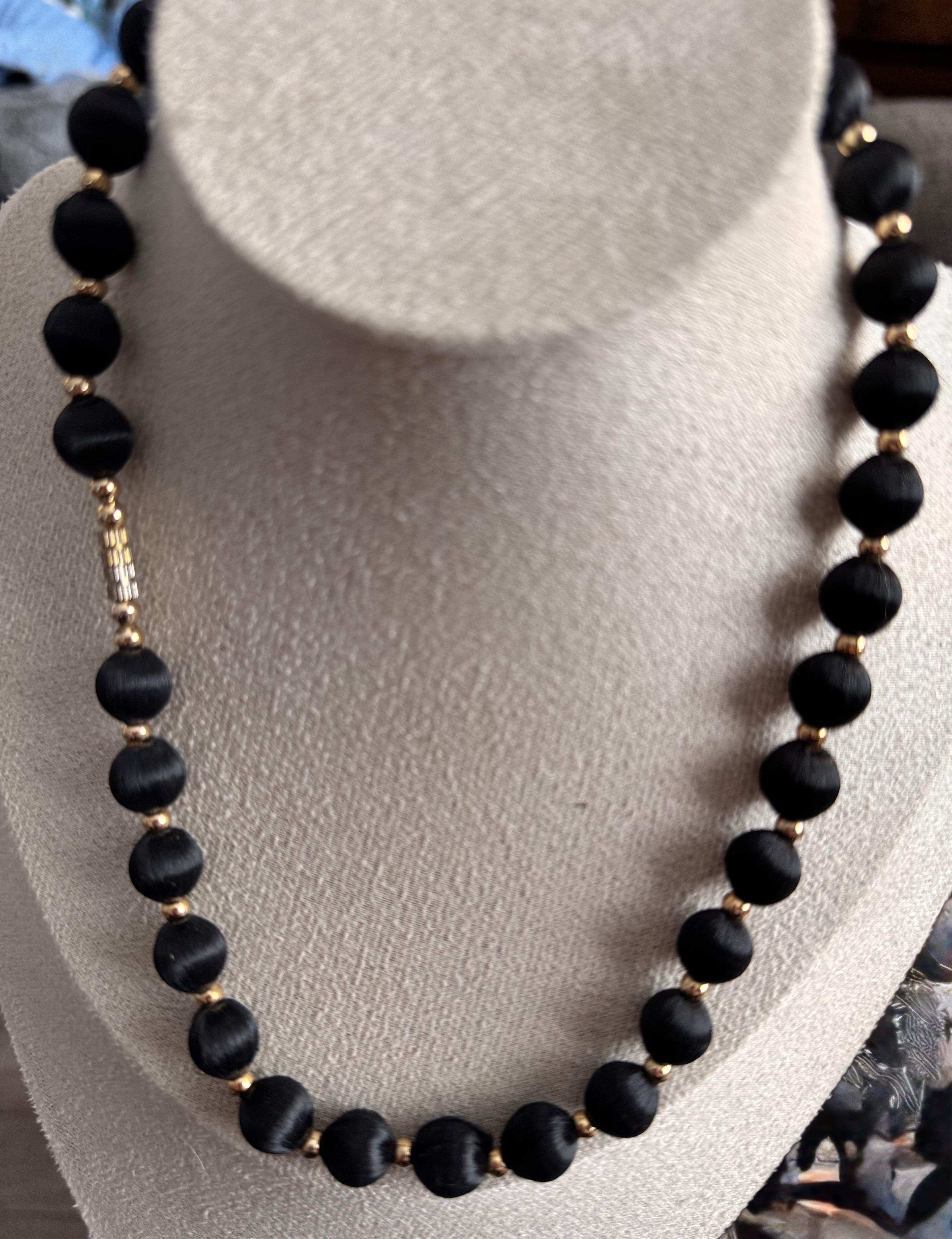 A black beaded necklace with small gold spacer beads displayed on a light gray fabric jewelry bust. Displaying the clasp type