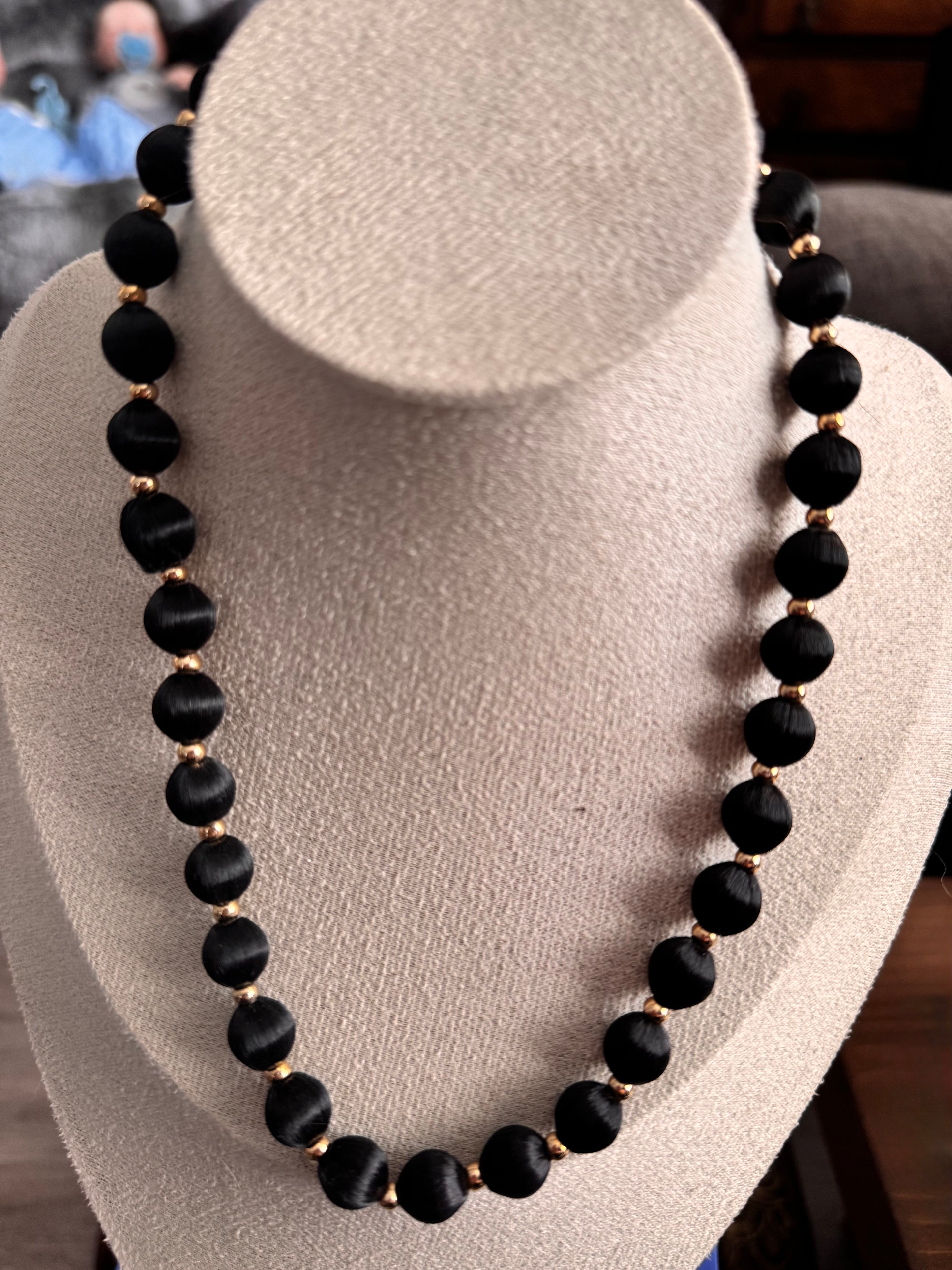 "A vintage beaded necklace with large round black satin-finish beads and small gold-toned spacer beads, displayed on a cream jewellery bust."