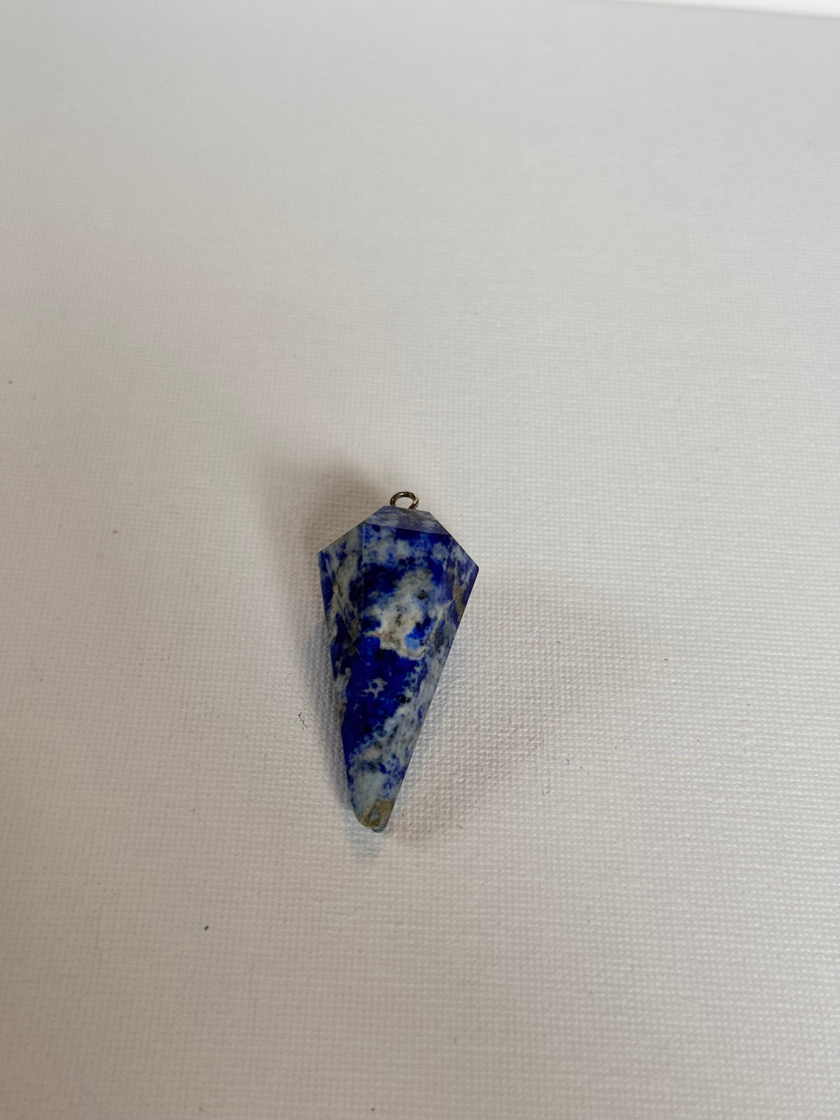A small pointed blue and white stone pendant with a tiny metal loop at the top, placed on a plain off-white textured surface.