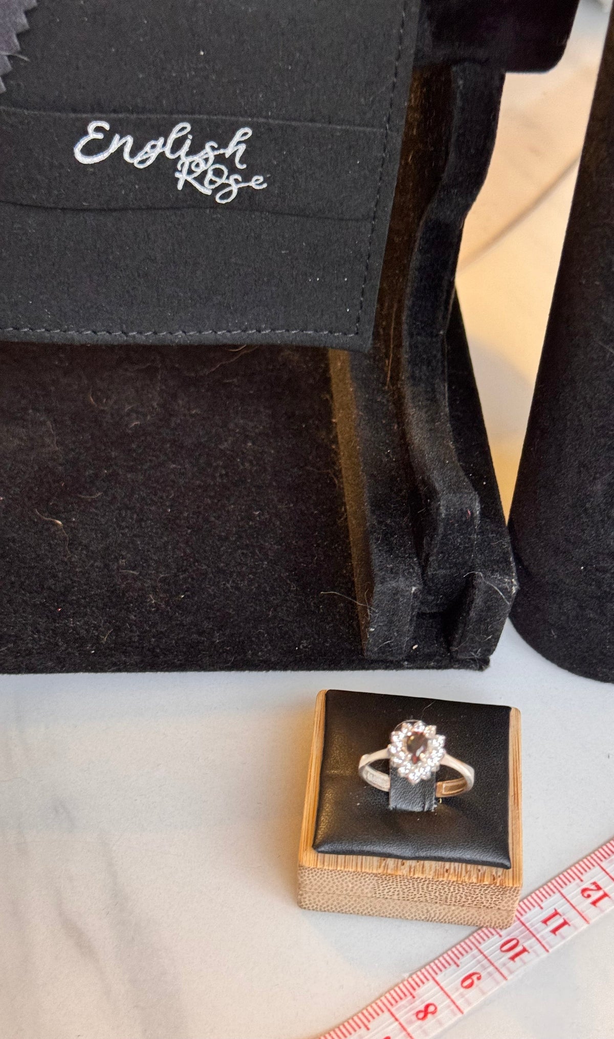 A silver ring with a pear-shaped halo ring. Moss agate centre stone and surrounded by cz's & is displayed on a small black jewelry box, with a measuring tape visible nearby and an 'English Rose' branded black velvet jewelry packaging & display in the background.