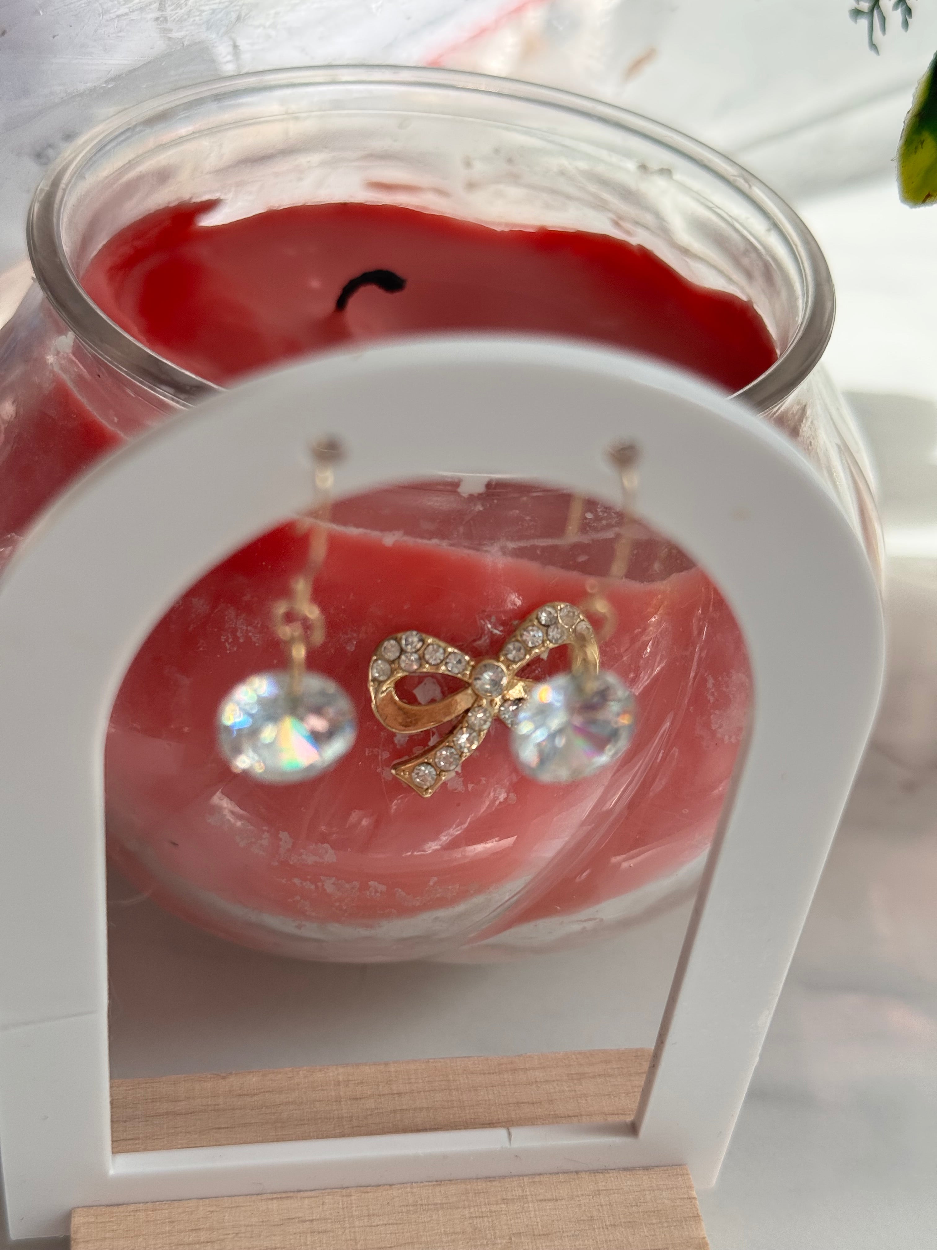A pair of dangling earrings displayed on a white stand in front of a red candle in a clear glass jar. The earrings feature 8mm diameter  round clear /Rainbow crystal drops hanging below.