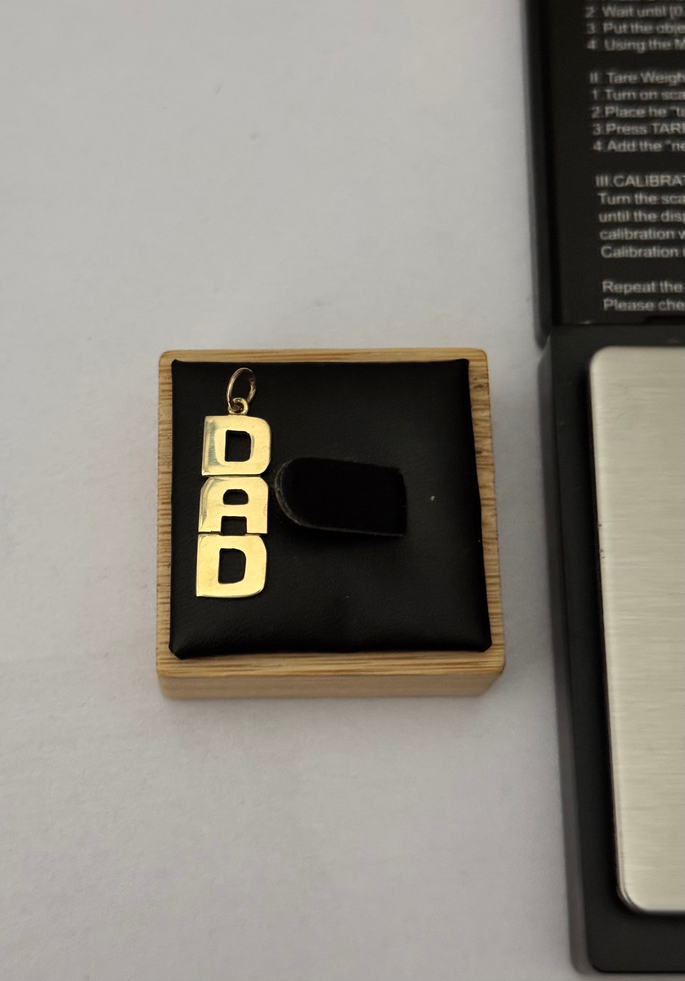A 9 ct hallmarked 'DAD' letter pendant charm displayed on a black velvet jewelry box, next to a black and silver device, on a light gray background.
