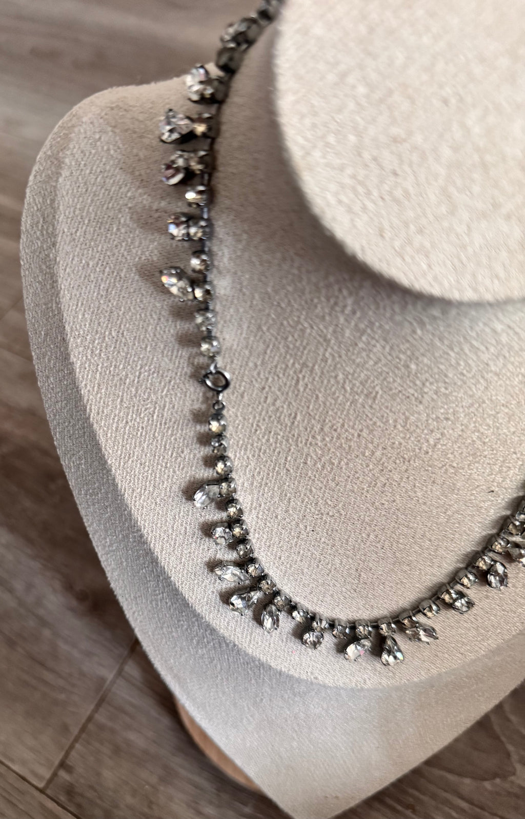 A close-up of a silver-toned necklace with clear teardrop-shaped crystals, displayed on a beige fabric jewelry bust against a wood floor background. Displaying the clasp too.