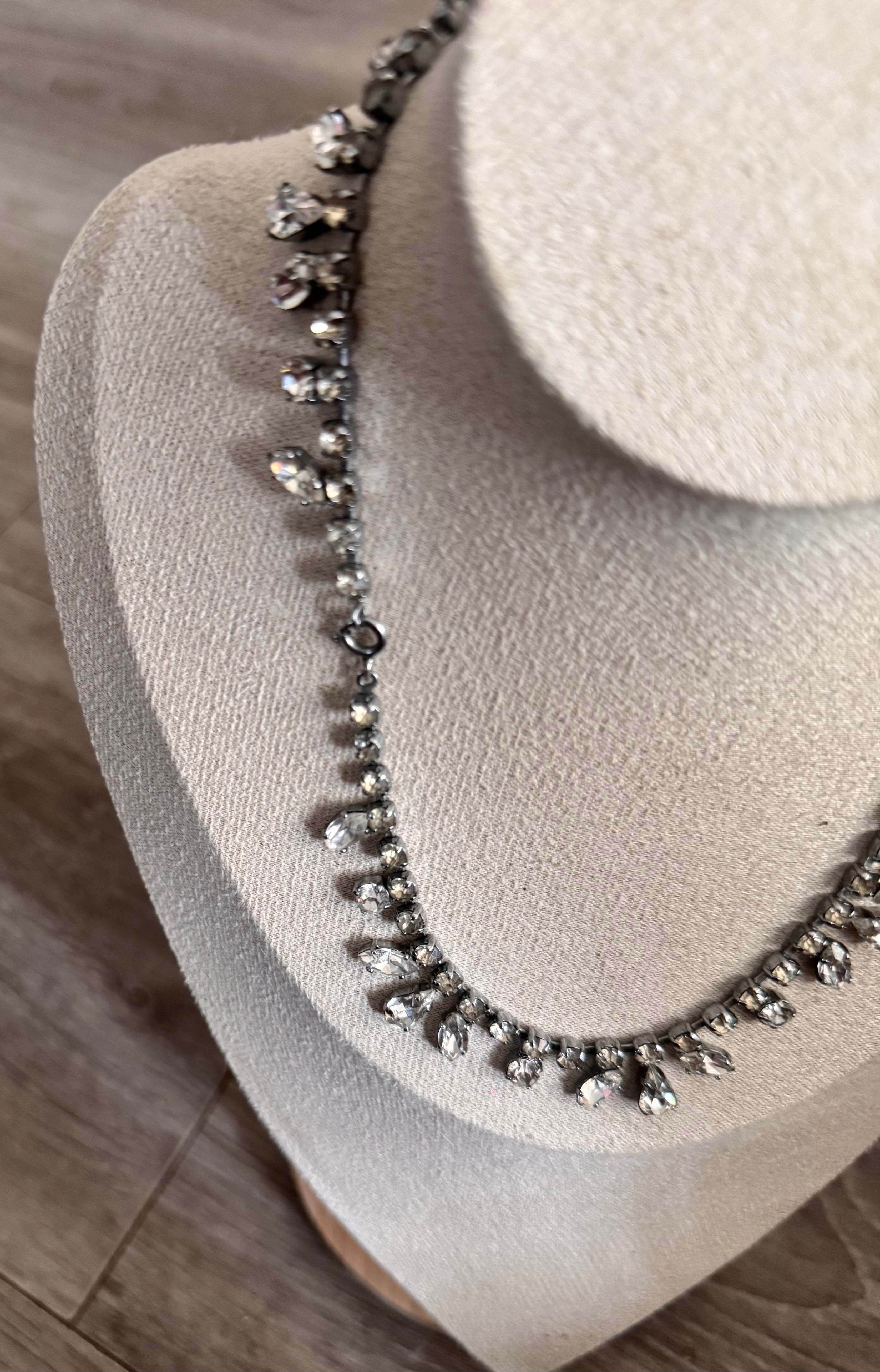 A close-up of a silver-toned necklace with clear teardrop-shaped crystals, displayed on a beige fabric jewelry bust against a wood floor background. Displaying the clasp too.